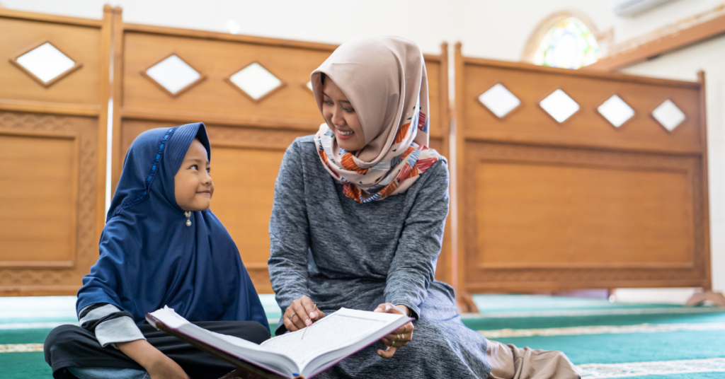 Female Quran teacher for children sitting with a young girl in hijab reading the Quran together in a mosque